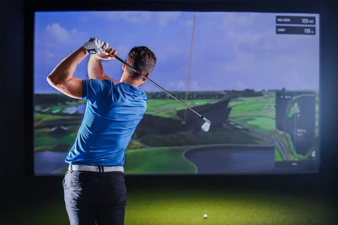 Golfer at the top of his backswing in front of a simulator screen showing TrackMan ball speed and carry